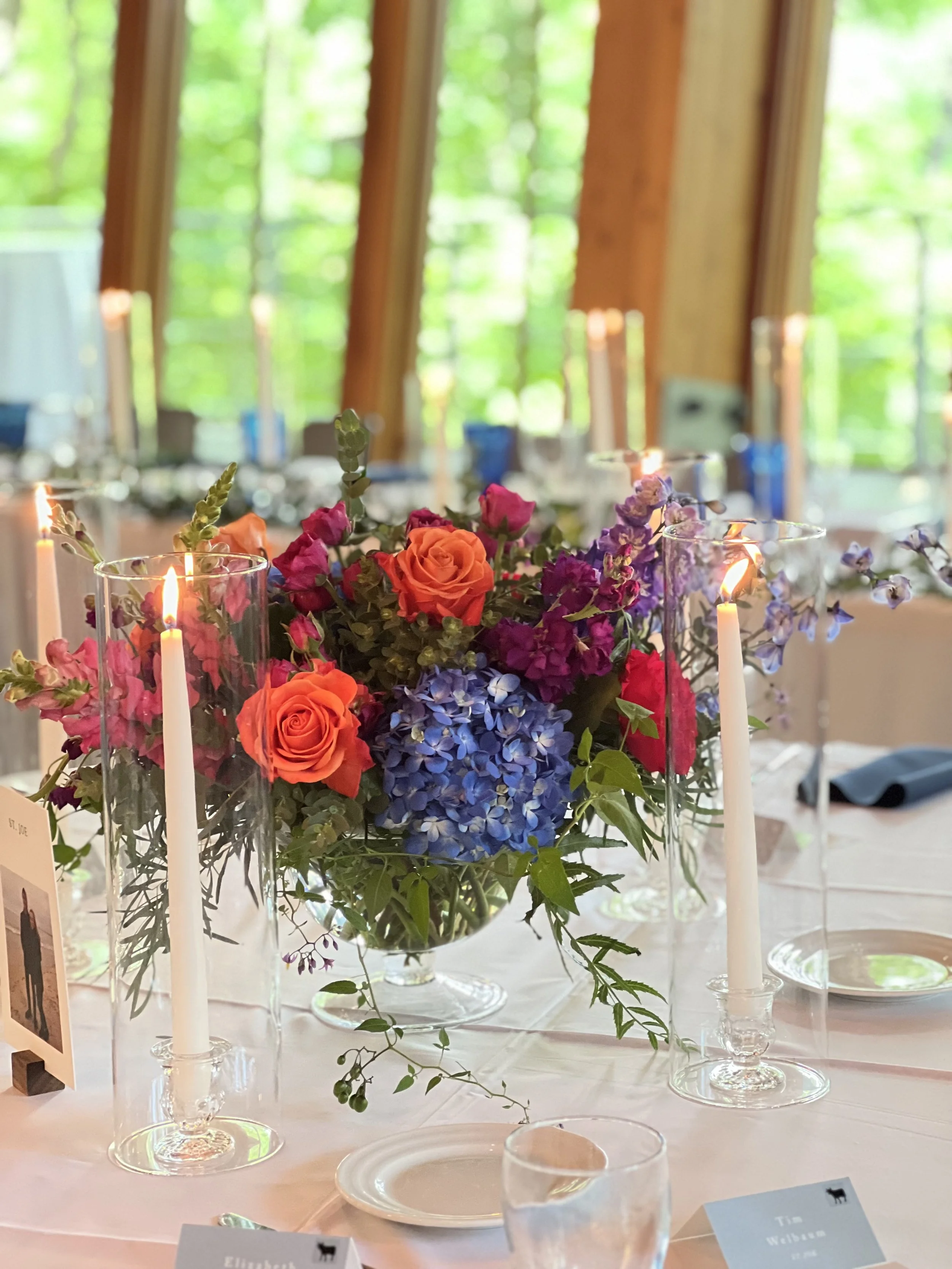 A compote centerpiece with orange, maroon and blue flowers